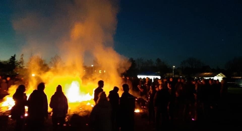 Nachtbild eines Osterfeuers, umgeben von einer Menschenmenge, deren Silhouetten sich gegen die hellen Flammen und den dunklen Himmel abzeichnen. Die Szene vermittelt eine Gemeinschaftsatmosphäre.