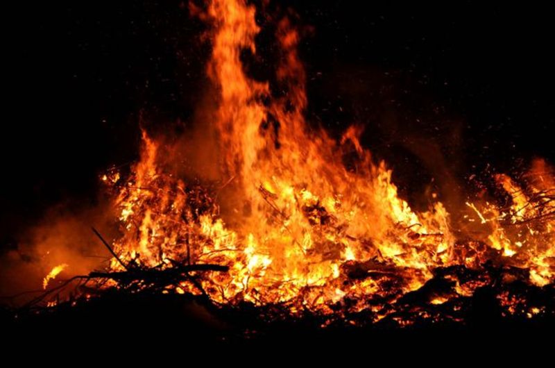 Ein intensives Feuer bei Nacht, das eine starke Glut und hohe Flammen aus Ästen und Holz entfacht, wobei die lebhaften Orange- und Gelbtöne stark im Kontrast zur dunklen Umgebung stehen.